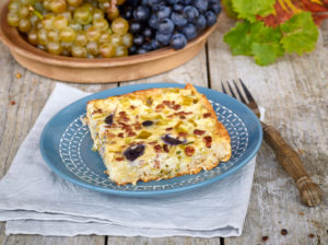 Quiche with grapes and leek, blue country plate on linen cloth, grapes in bowl, fork with wooden handle, table with untreated table top Quiche with grapes and leek, blue country plate on linen cloth, grapes in bowl, fork with wooden handle, table with untreated table top